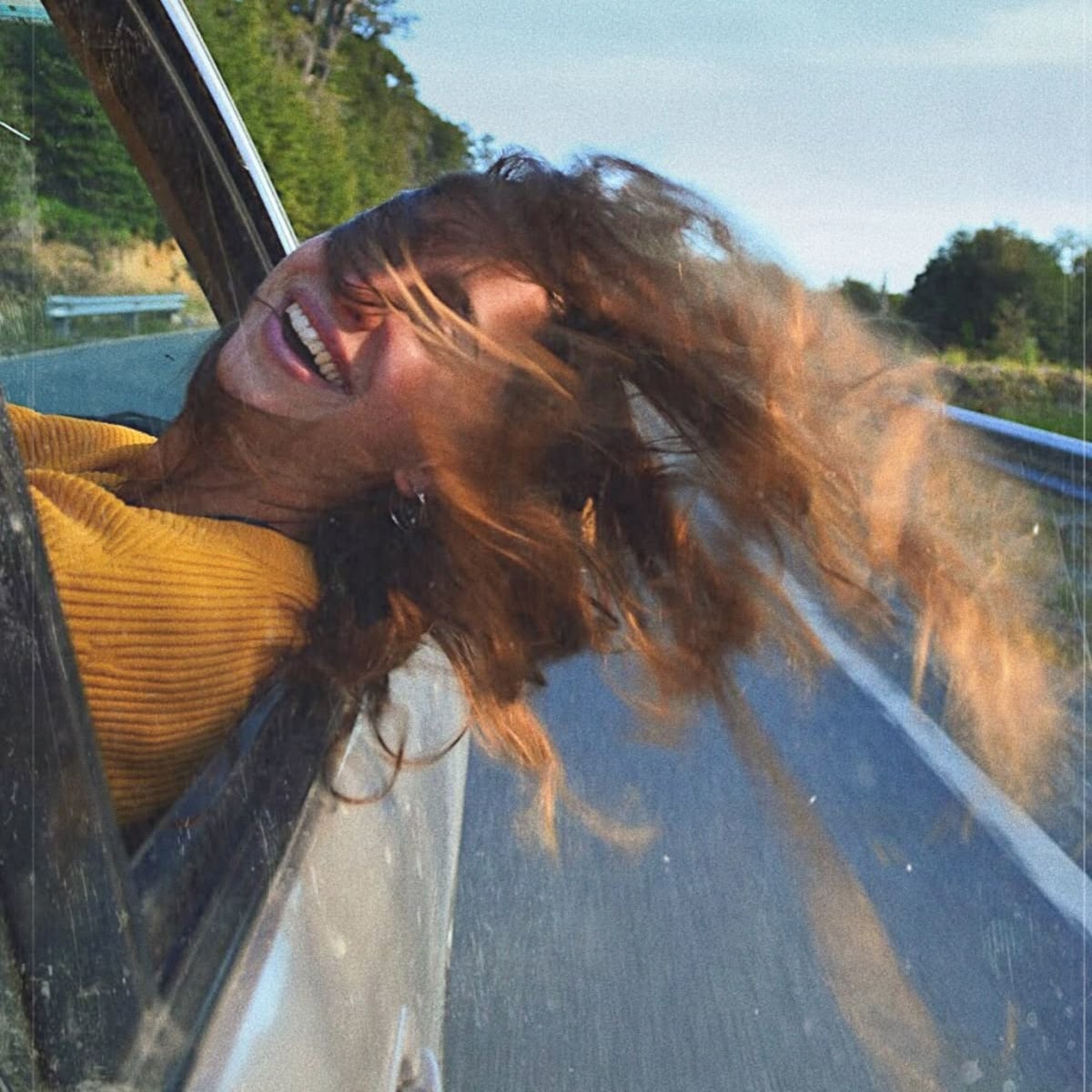 A person with long, wavy hair leans out of a moving car window, smiling joyfully as their hair blows in the wind. They are wearing a yellow sweater, and the car is driving down a road bordered by greenery.