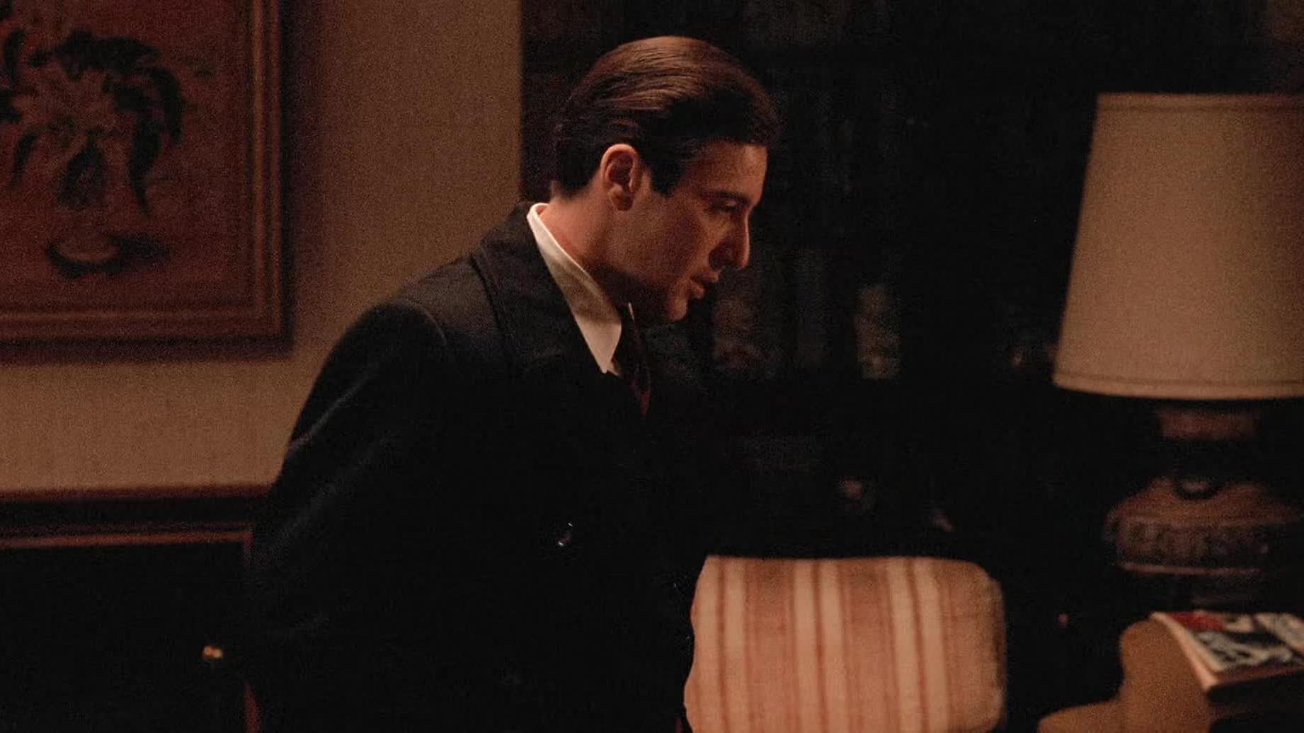 A man in a dark suit stands indoors, looking serious and facing left. Behind him are a striped armchair, a table with magazines, a lamp, and a framed artwork on the wall. The room is dimly lit.