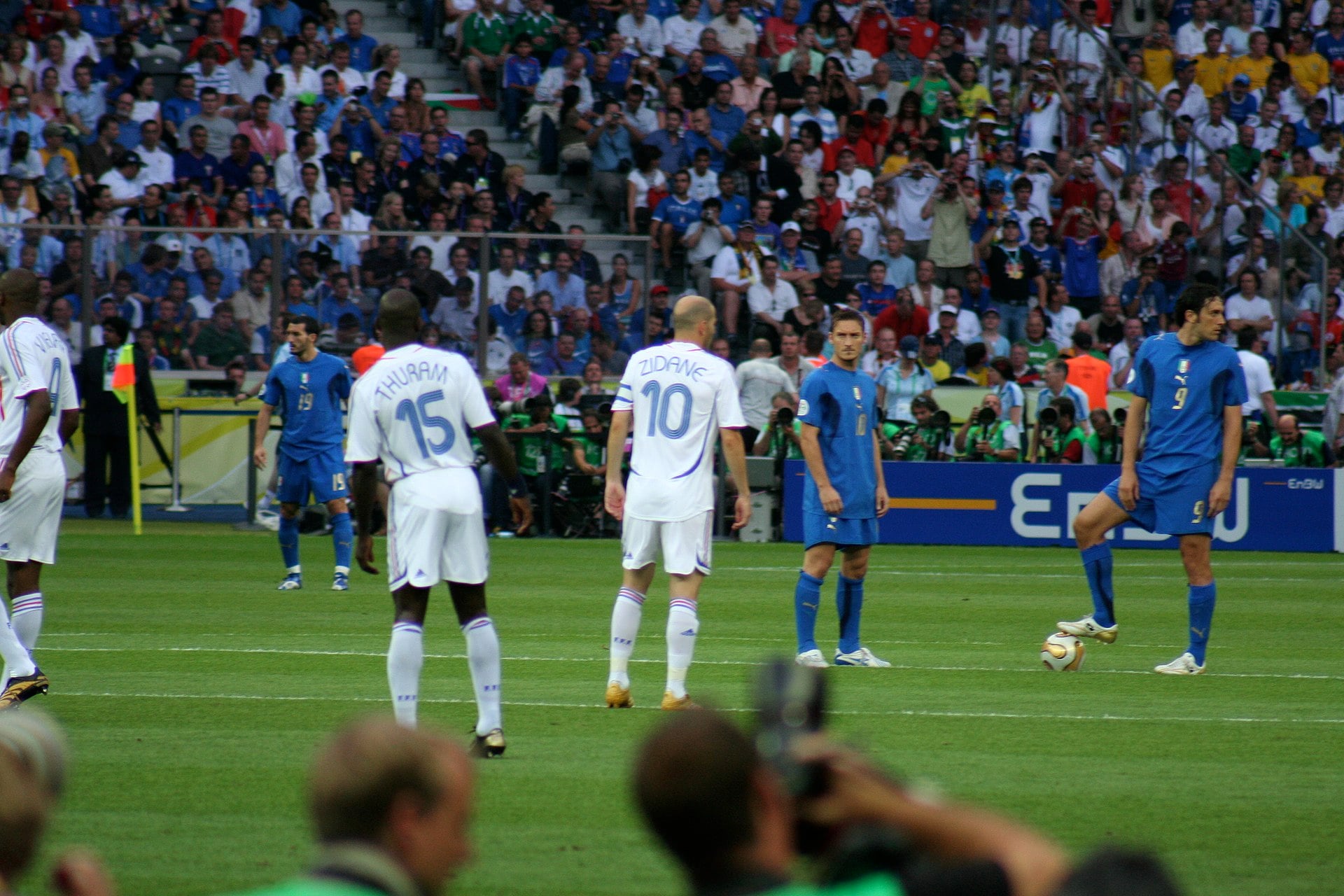 A soccer match in a crowded stadium shows players in white and blue jerseys ready for play, with a player in blue about to kick the ball as spectators watch from the stands.