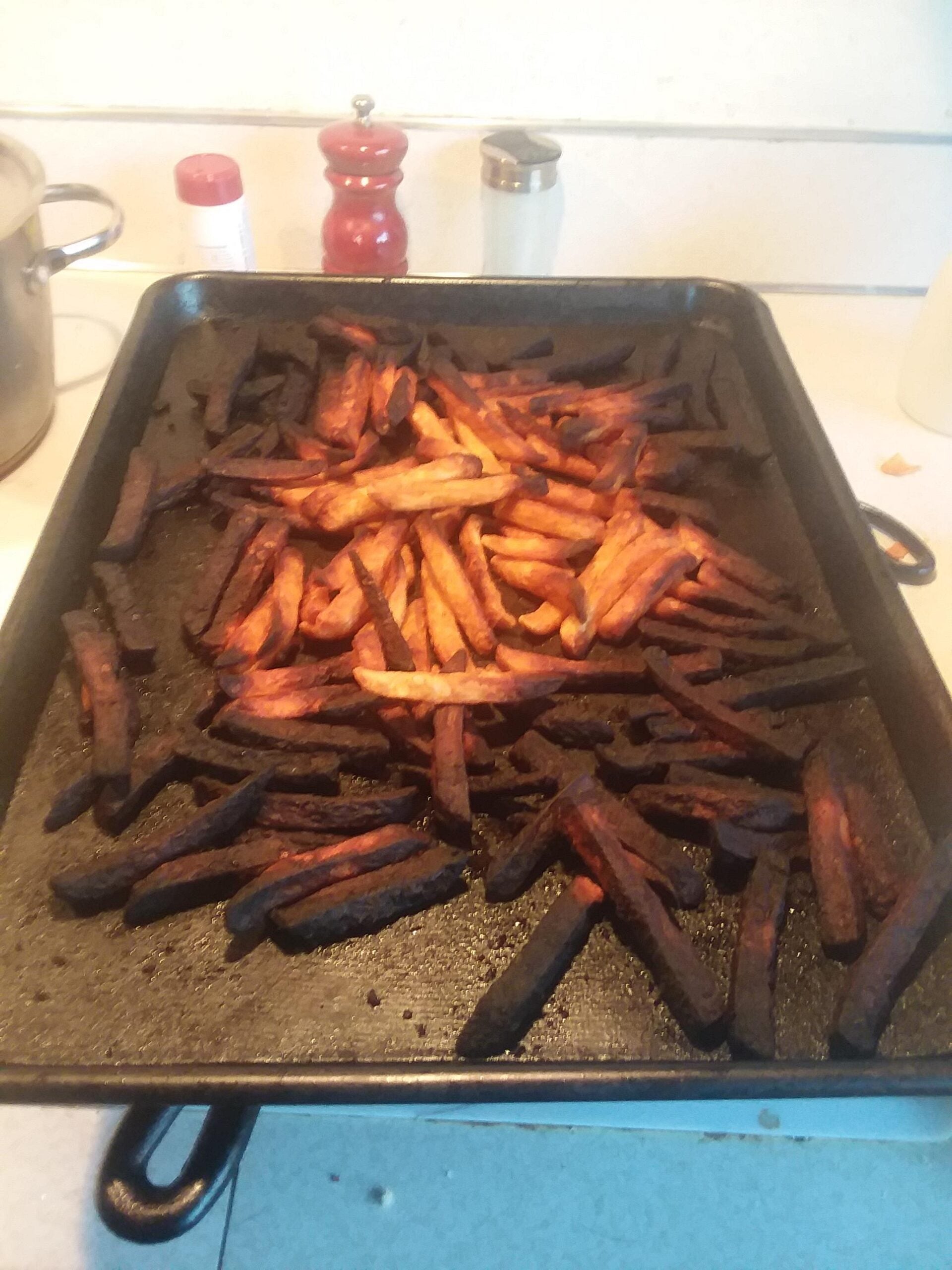 A baking tray of homemade fries, many of which are burnt and dark brown, sits on a stovetop. In the background are a red pepper grinder, salt shaker, and a metal pot.
