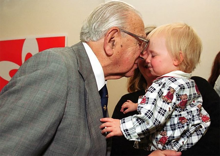 An elderly man in a suit gently touches foreheads with a young child, who is being held by an adult. Both appear to be sharing a tender, affectionate moment. A red and white design is visible in the background.