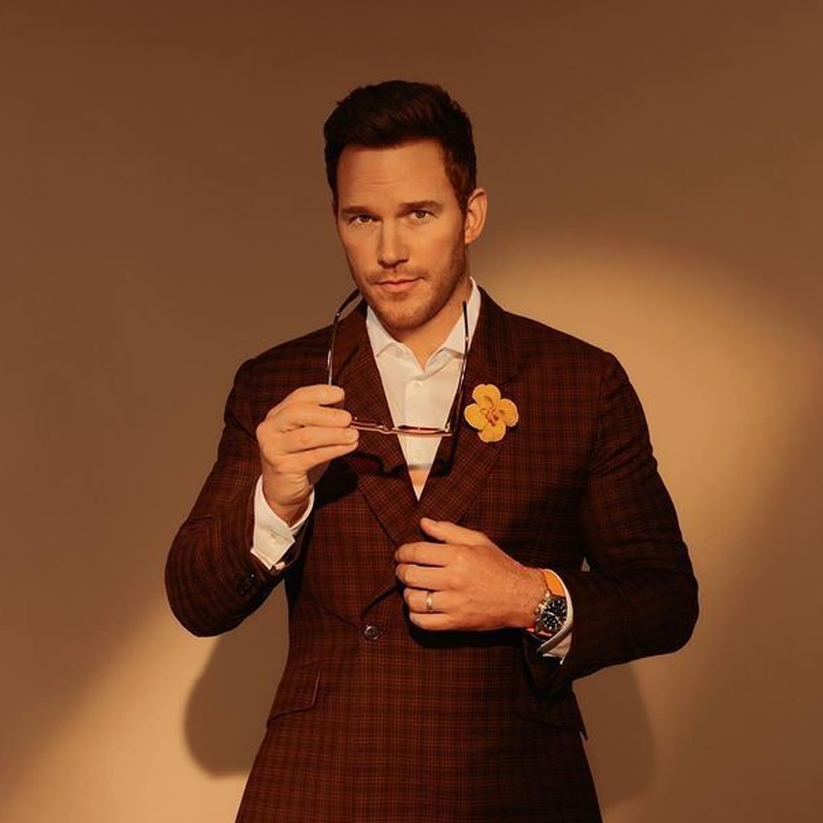 A man in a brown plaid suit with a white shirt and a yellow flower pin holds sunglasses and looks at the camera. He stands against a beige background in warm lighting.