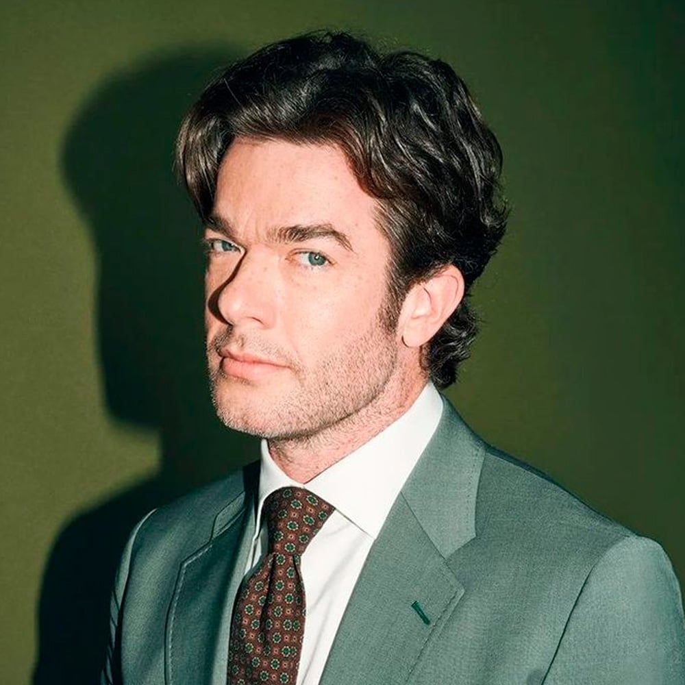 A man with light skin, dark hair, and blue eyes wears a gray suit, white shirt, and patterned tie. He stands against a green background, facing slightly left, with a serious expression.
