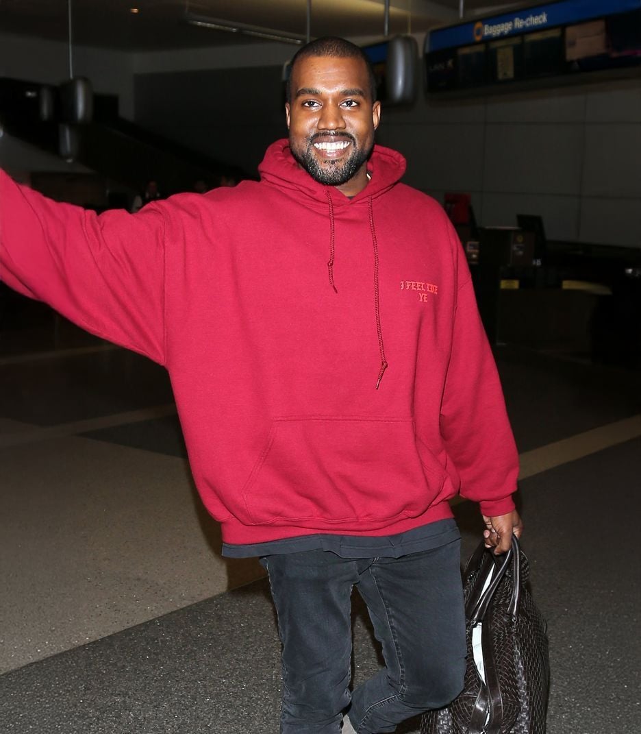 A man wearing a red hoodie, black pants, and carrying a textured bag, smiles and waves while walking through an airport terminal.
