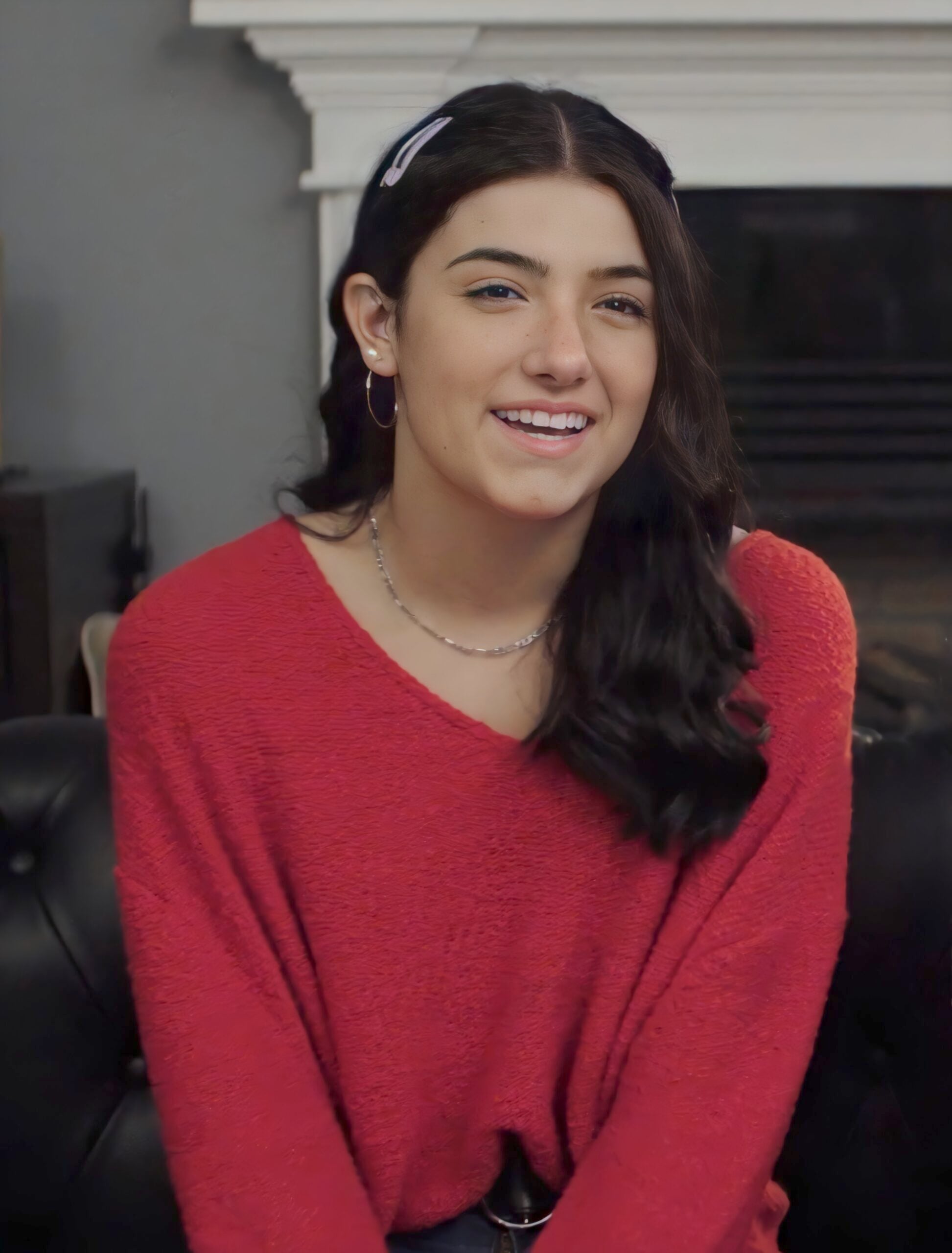 A young woman with long dark hair, wearing a red sweater and a hair clip, smiles while sitting on a black couch in a cozy living room.