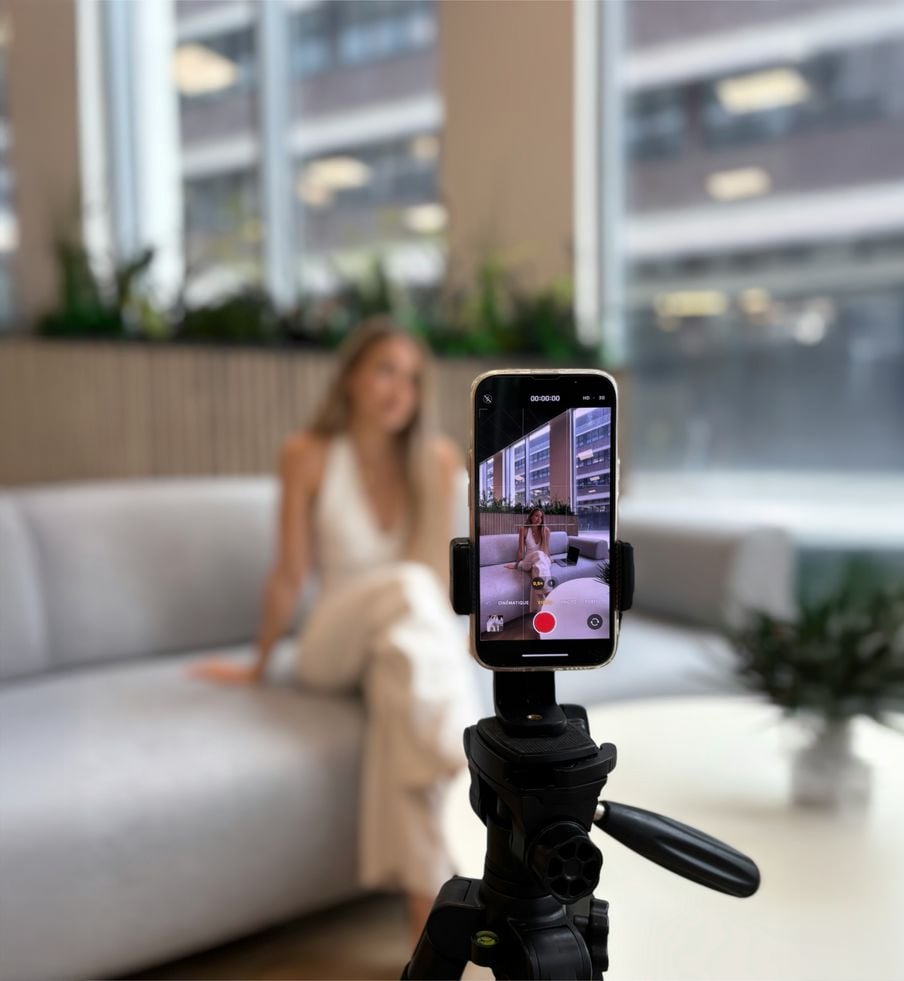 A smartphone on a tripod records a woman in a white outfit sitting on a gray couch in a modern, bright room with large windows and plants.