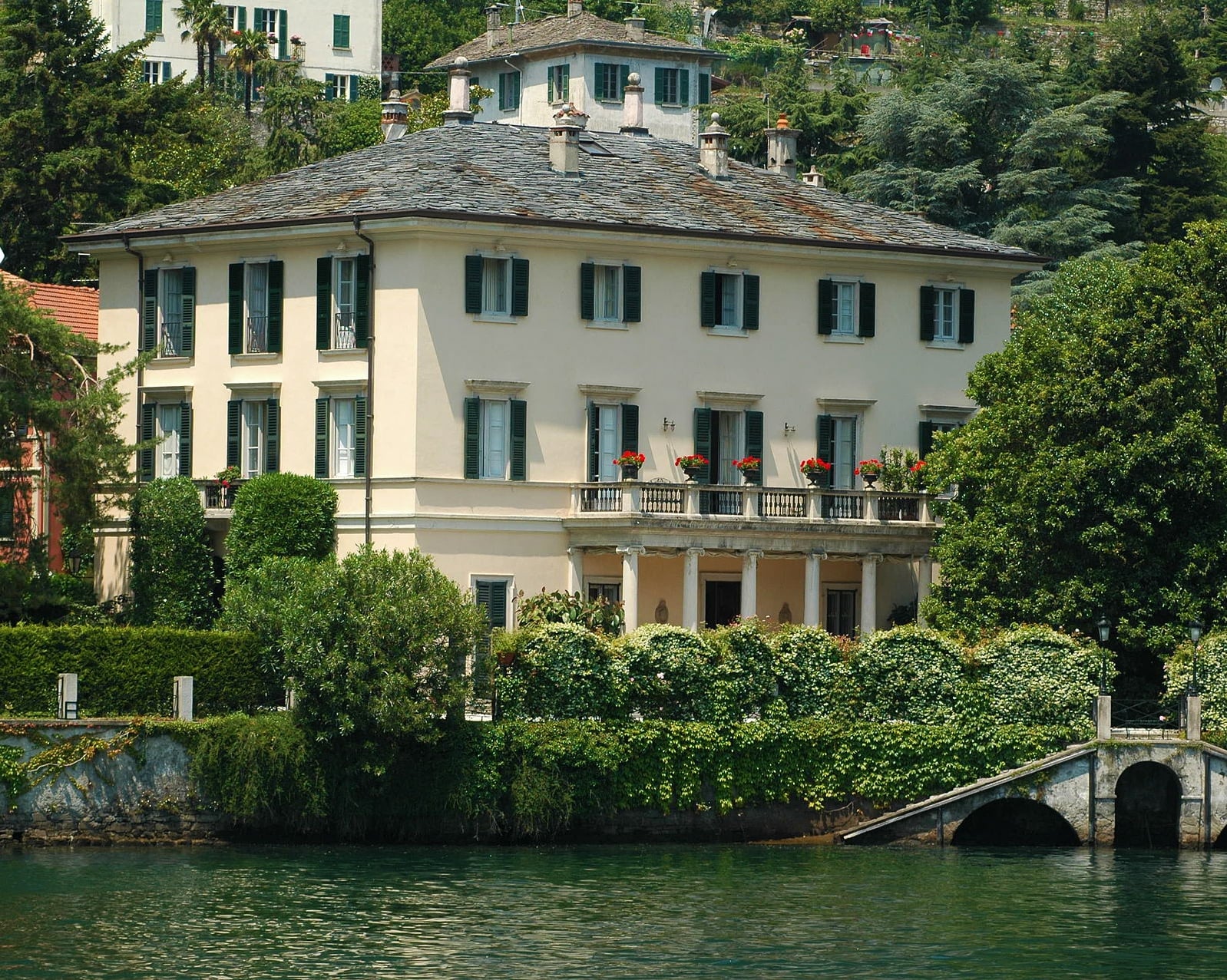 Large, elegant cream-colored villa with green shutters and a slate roof, surrounded by lush greenery and trees, sits beside a calm body of water, with an arched stone dock at the shore.