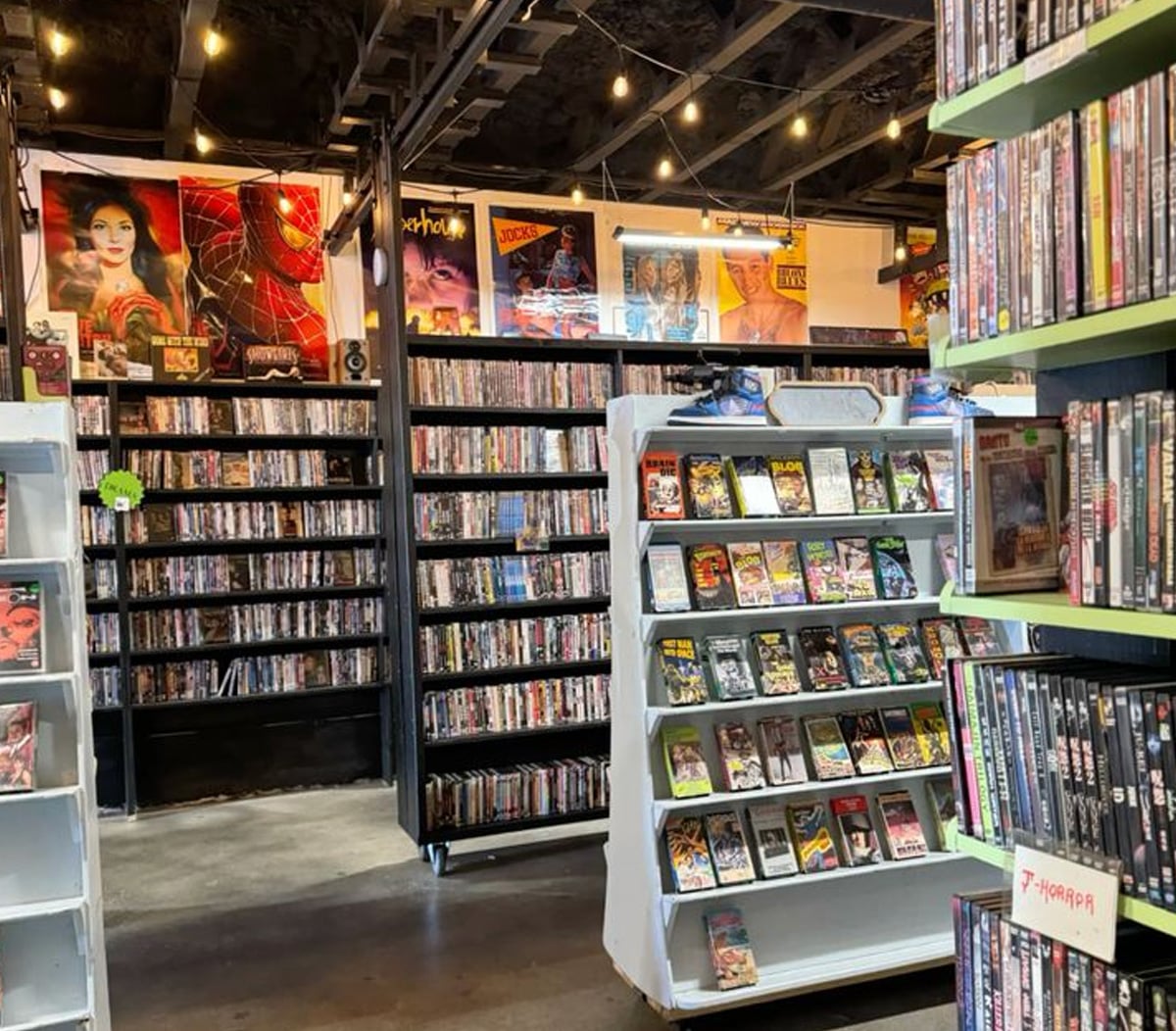 A video rental store with shelves full of DVDs and VHS tapes, including a "Horror" section. Colorful movie posters and string lights decorate the space, creating a nostalgic retro atmosphere.