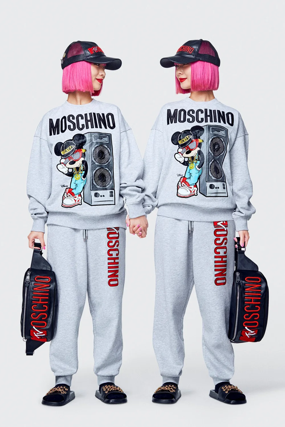 Two people with pink bob haircuts, wearing matching gray Moschino sweatshirts featuring Mickey Mouse, gray Moschino sweatpants, black slide sandals, black Moschino bags, and black caps, standing side by side holding hands.