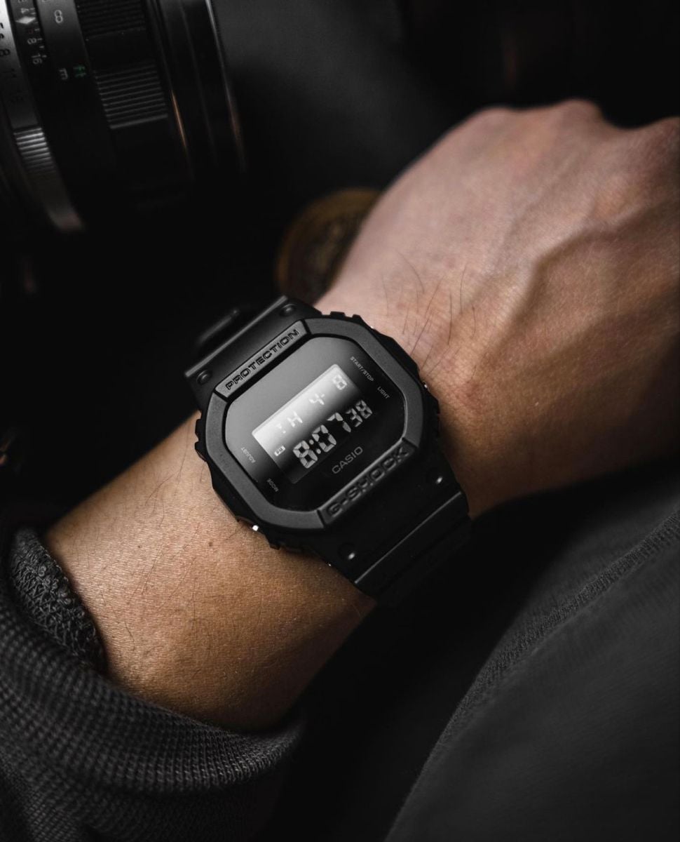 A close-up of a person’s wrist wearing a black Casio digital watch displaying the time 8:01:38. The person is dressed in dark clothing, and a camera lens is visible in the background.