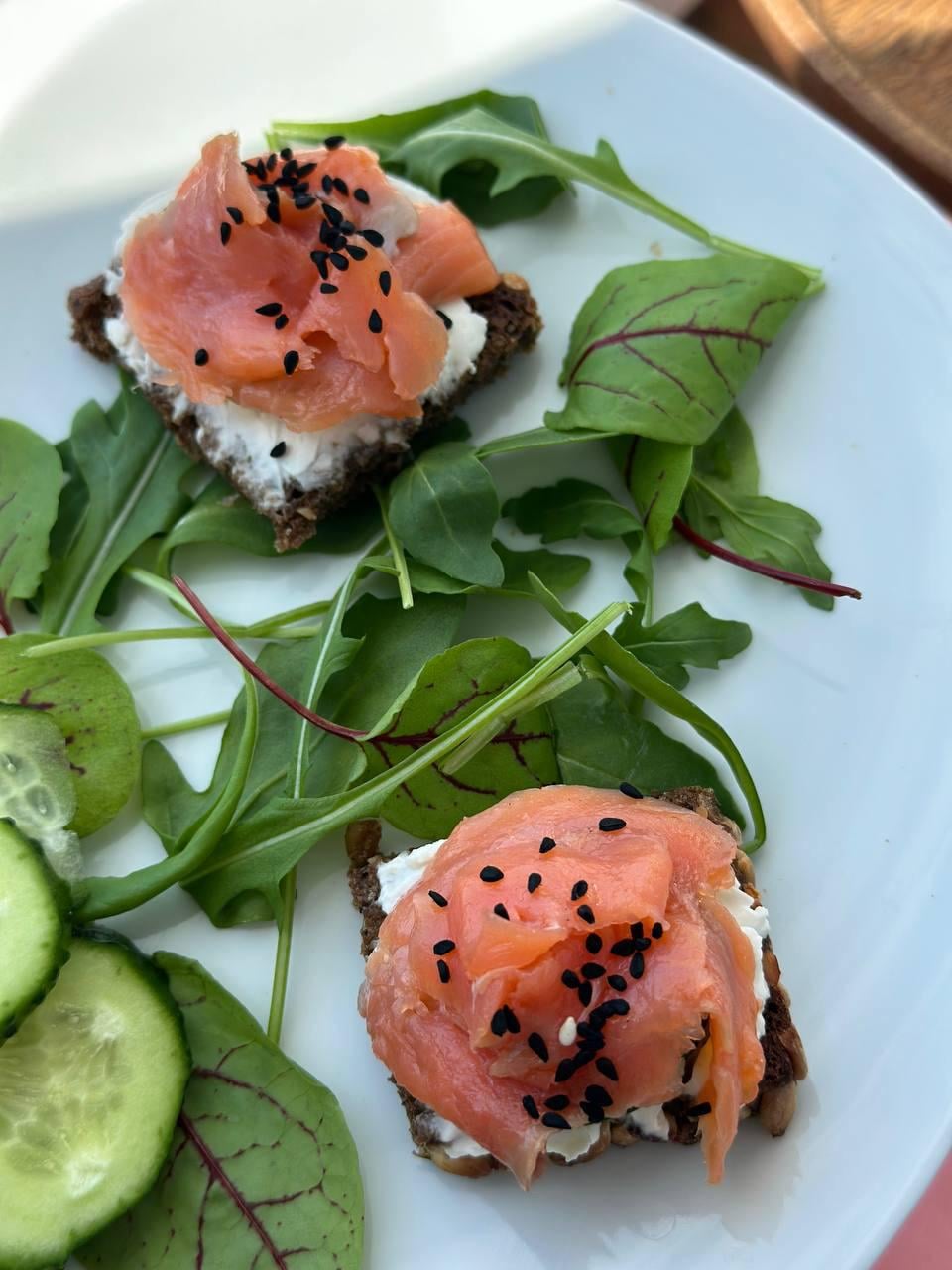 Two small pieces of dark bread topped with cream cheese, smoked salmon, and black sesame seeds, served on a white plate with fresh arugula, cucumber slices, and red-veined greens.