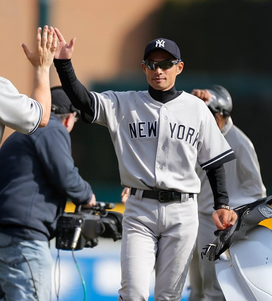 A New York Yankees baseball player in uniform and sunglasses gives a high five to a teammate, with a cameraman and another player visible in the background.