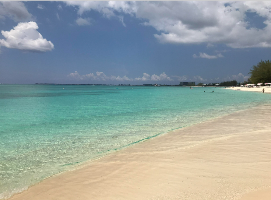 A tranquil beach scene with clear turquoise water gently washing onto soft, sandy shores under a partly cloudy sky. A few people are seen in the distance enjoying the calm ocean.