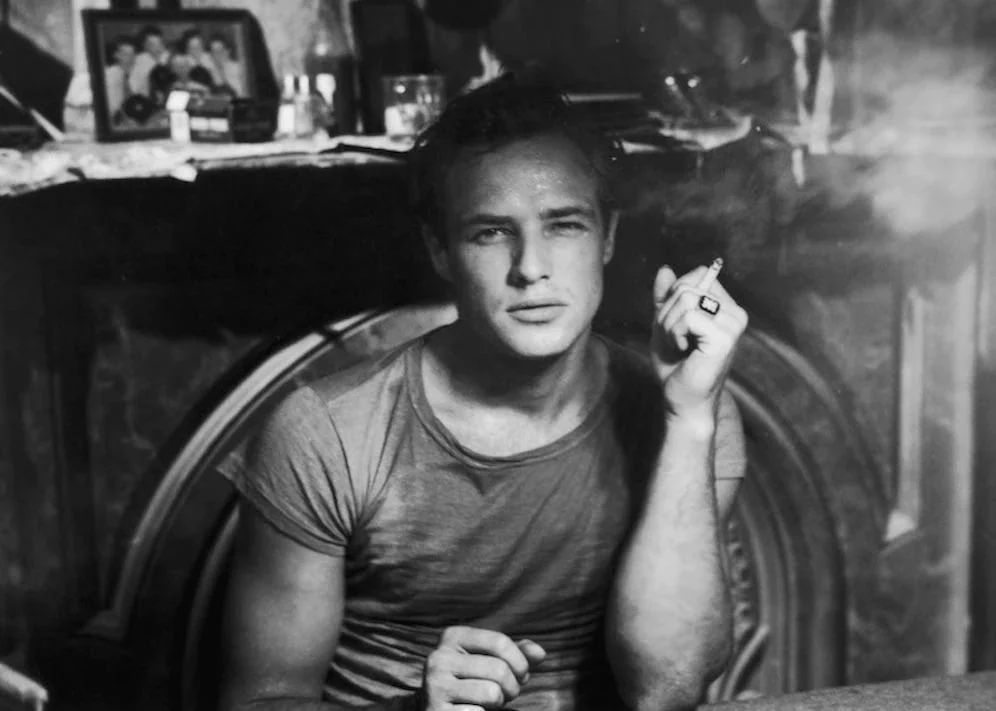 A young man in a fitted t-shirt sits in front of a fireplace, holding a cigarette. Family photos and small objects are on the mantle behind him. The scene is in black and white, giving it a classic, vintage look.