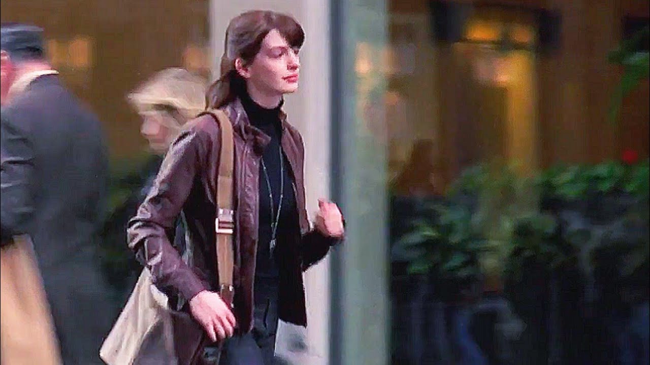 A woman with brown hair walks outside in a city wearing a brown leather jacket and carrying a tan shoulder bag. People and greenery are blurred in the background.
