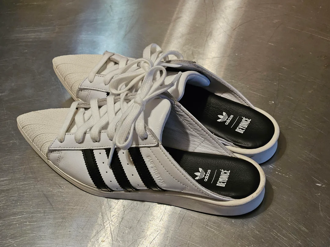 A pair of white Adidas shoes with black stripes are styled as open-back slip-ons, placed on a metallic surface. The insoles feature the Adidas logo and the word "BY WONG.
