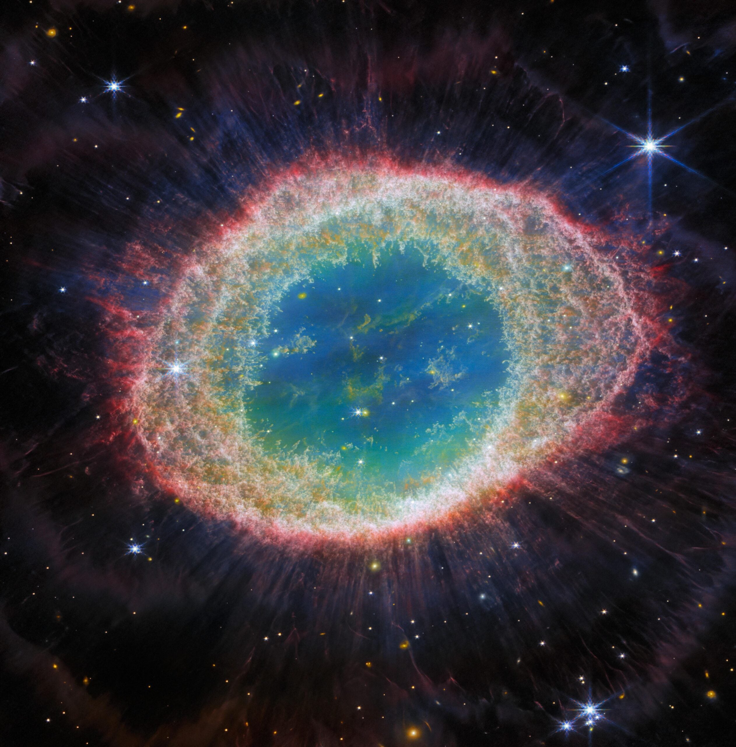 A vibrant, colorful nebula shaped like a glowing ring in space, with a bright blue-green center, surrounded by layers of red, pink, and white gas, set against a background of stars.