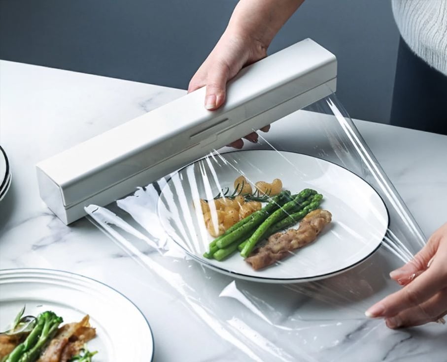 A person covers a plate of food, including asparagus and chicken, with clear plastic wrap using a white dispenser on a marble countertop.
