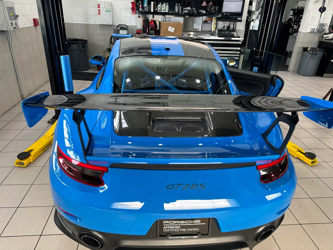 A bright blue Porsche GT2 RS with a large rear wing is parked in a clean automotive workshop, with its doors open and yellow lift arms nearby. Equipment and tools are visible in the background.
