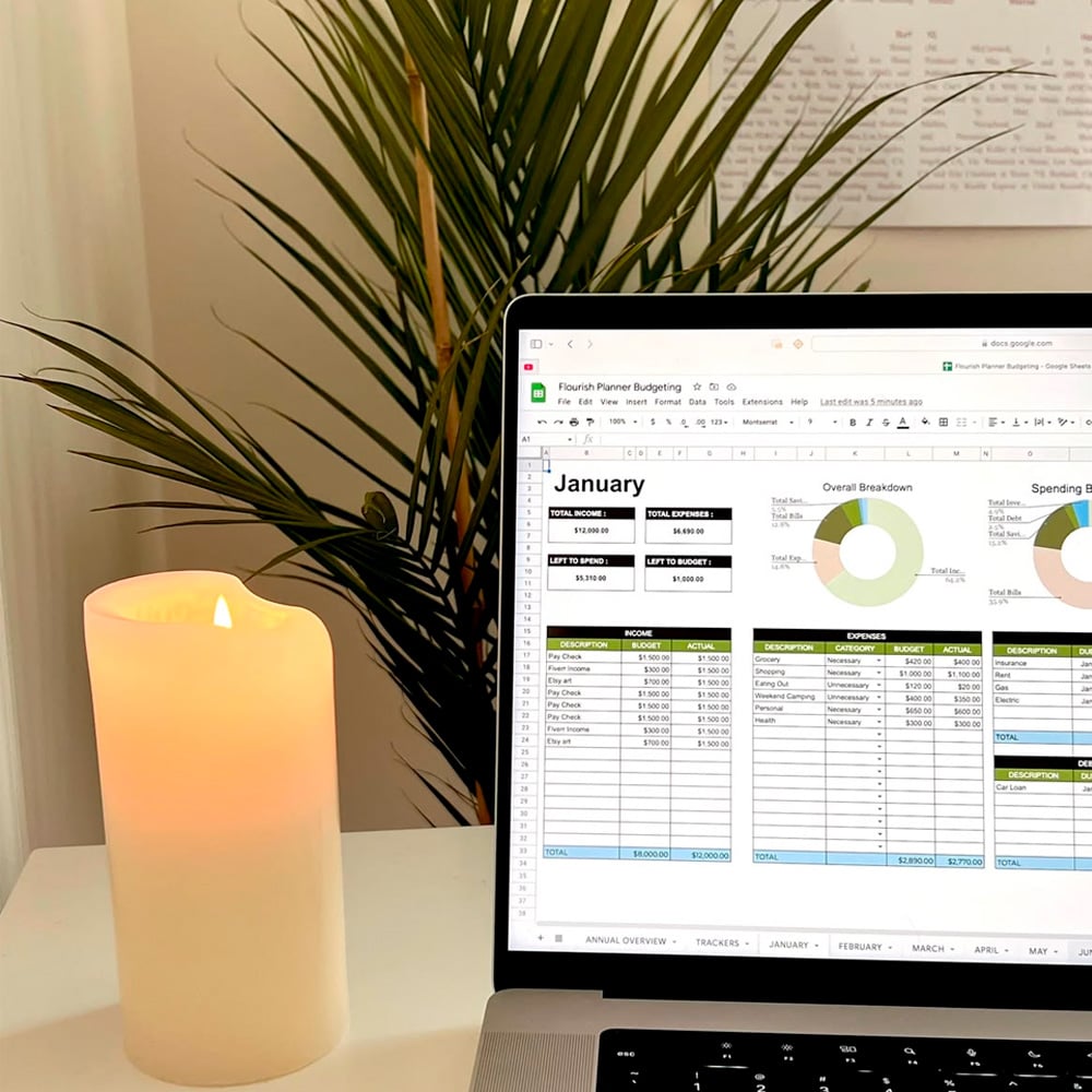 A lit white candle sits beside an open laptop displaying a budgeting spreadsheet for January. A green plant and a whiteboard with writing are visible in the background.