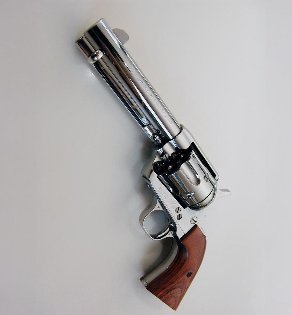 A silver revolver with a wooden grip is displayed on a light gray surface, angled diagonally with the barrel pointing upward and to the left.