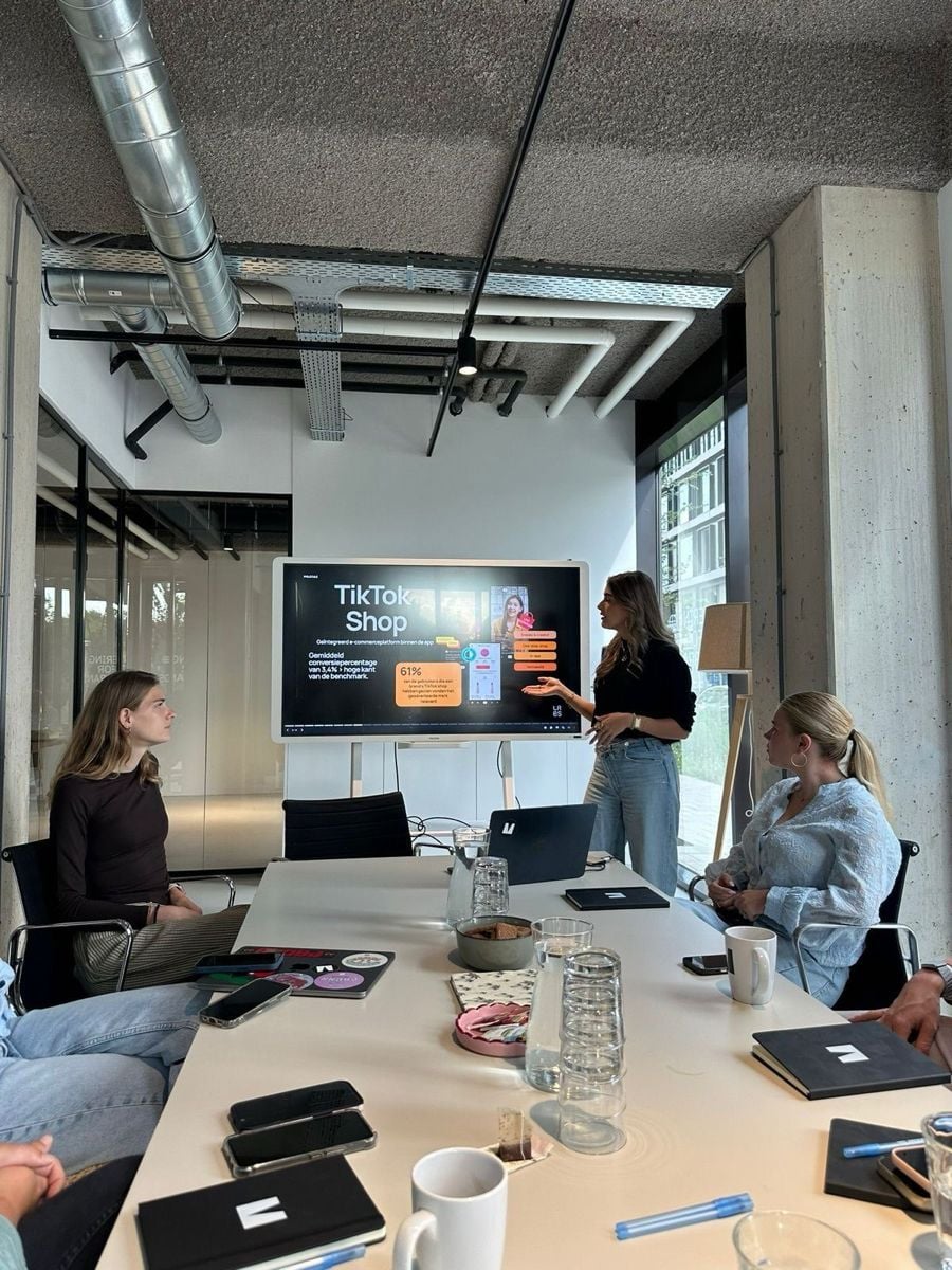 Four people sit around a conference table while a woman stands and presents information about TikTok Shop on a large screen. Laptops, notebooks, and drinks are on the table in a modern office setting.