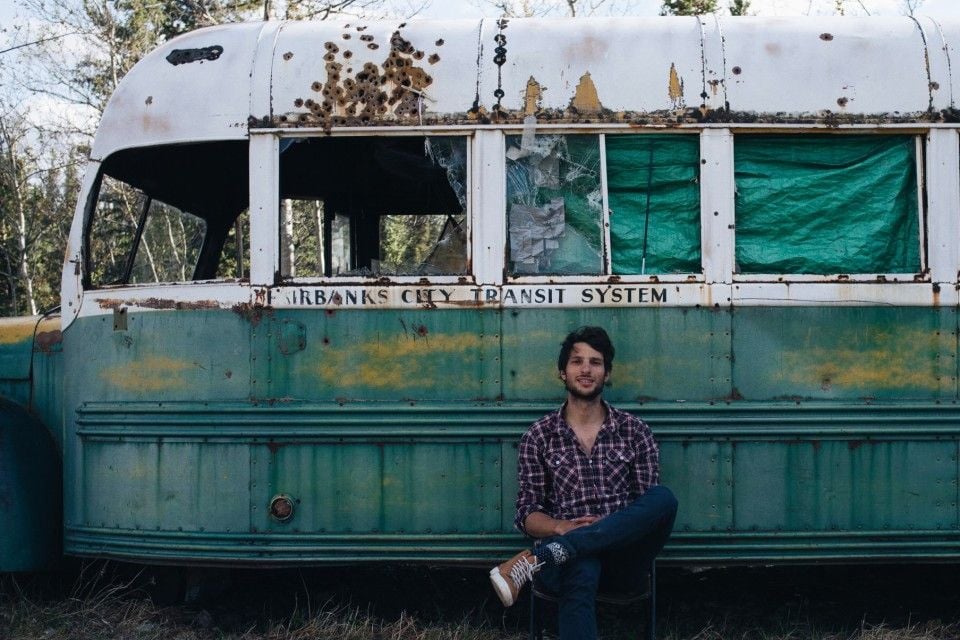 A man in a plaid shirt sits on a chair in front of an old, weathered bus with broken windows and faded paint. The bus has "Fairbanks City Transit System" written on its side.