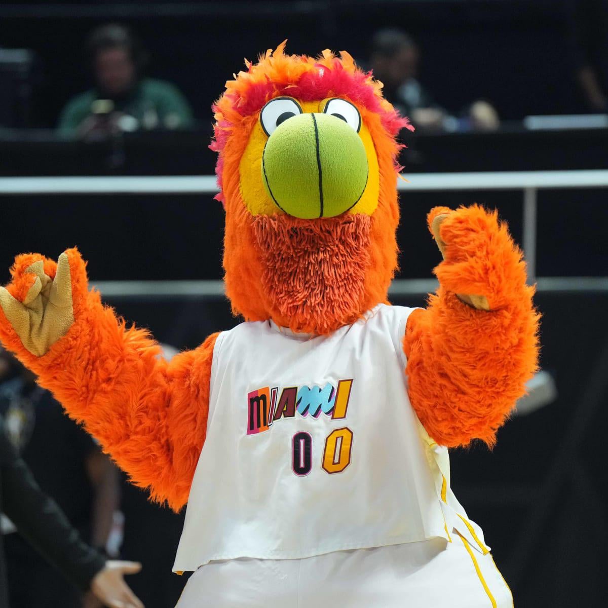 An orange, fuzzy bird mascot wearing a white "Miami 00" jersey stands on a basketball court with both hands raised, appearing to cheer or dance.