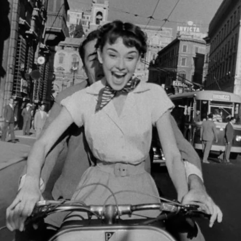 A young woman with short hair and a scarf around her neck smiles excitedly while riding a scooter through a busy city street, with a man seated behind her. The scene is in black and white.
