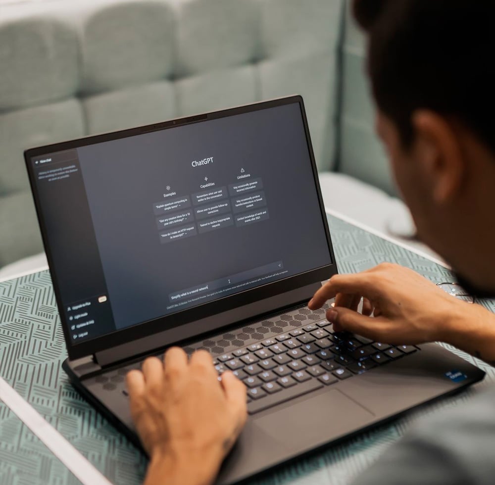 A person types on a laptop displaying the ChatGPT interface on the screen, with blurred hands on the keyboard and a light patterned desk surface beneath the device.