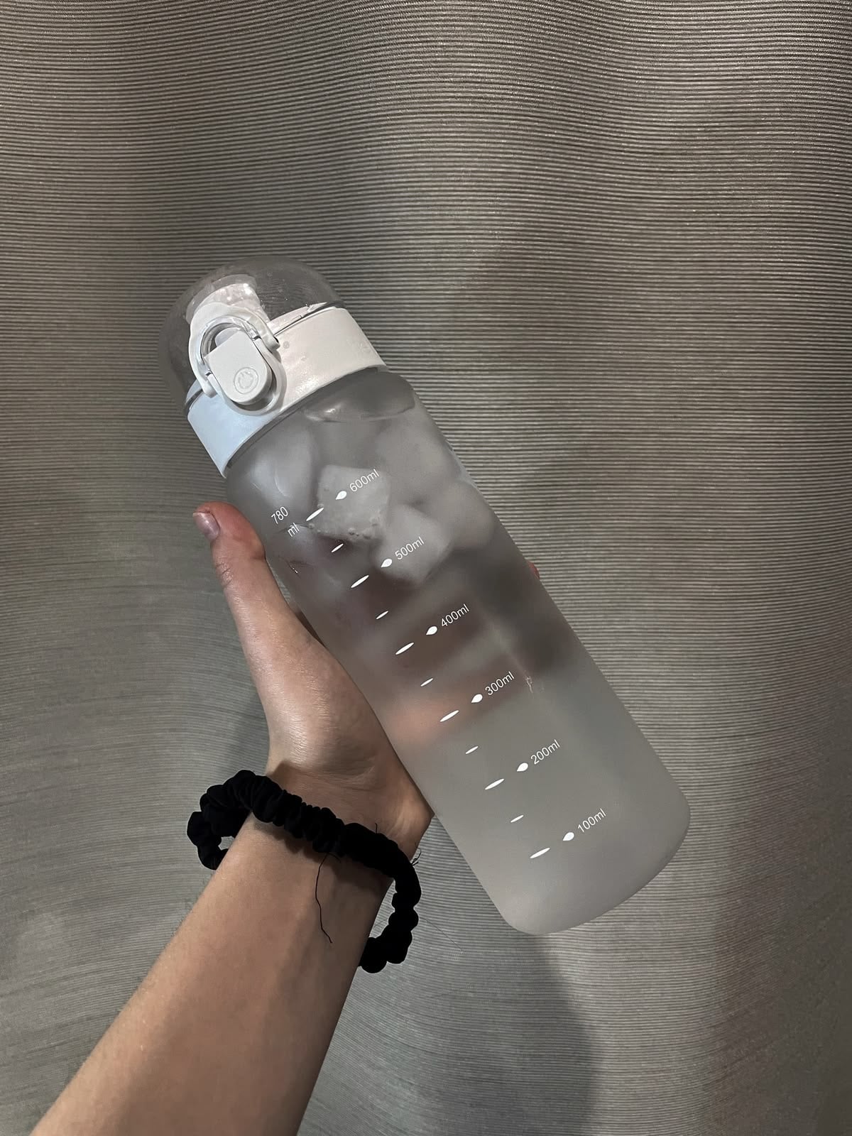 A hand holding a frosted water bottle with measurement markings and filled with ice. The person is wearing a black scrunchie on their wrist, and the background is a textured gray surface.