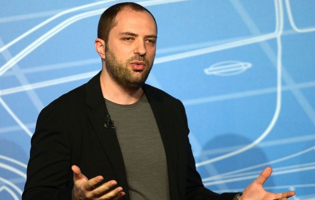 A man with short hair and a beard, wearing a black blazer and gray shirt, gestures with his hands while speaking in front of a blue background with white geometric line patterns.