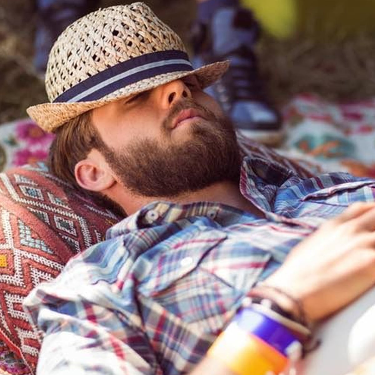 A bearded man in a plaid shirt lies on a patterned pillow outdoors, wearing a straw hat tilted over his eyes. He has colorful bracelets on his wrist and appears to be resting or napping.