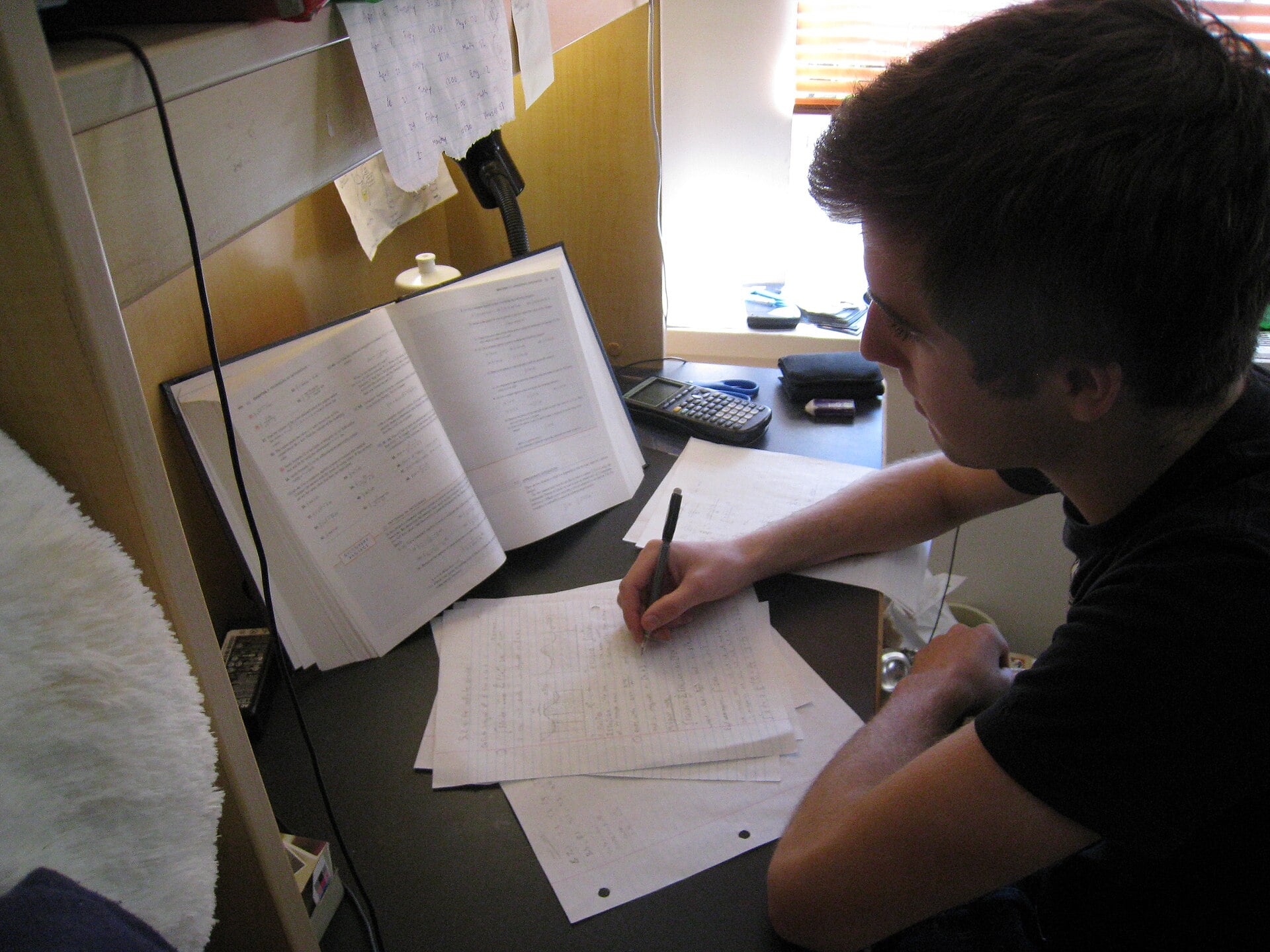 A person sits at a desk with an open textbook, writing on sheets of paper. The desk has a calculator, lamp, and various papers, suggesting they are studying or doing homework. Light comes in through a nearby window.