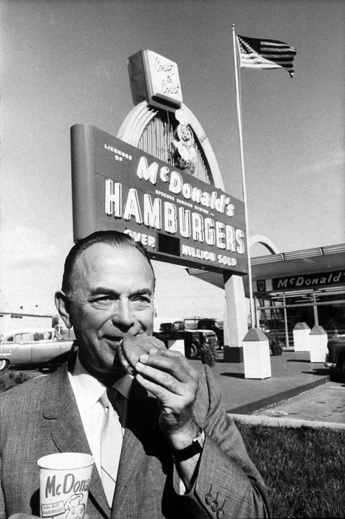 A man in a suit holds a McDonald’s cup and eats a hamburger in front of a vintage McDonald’s restaurant with a large sign and a waving American flag.