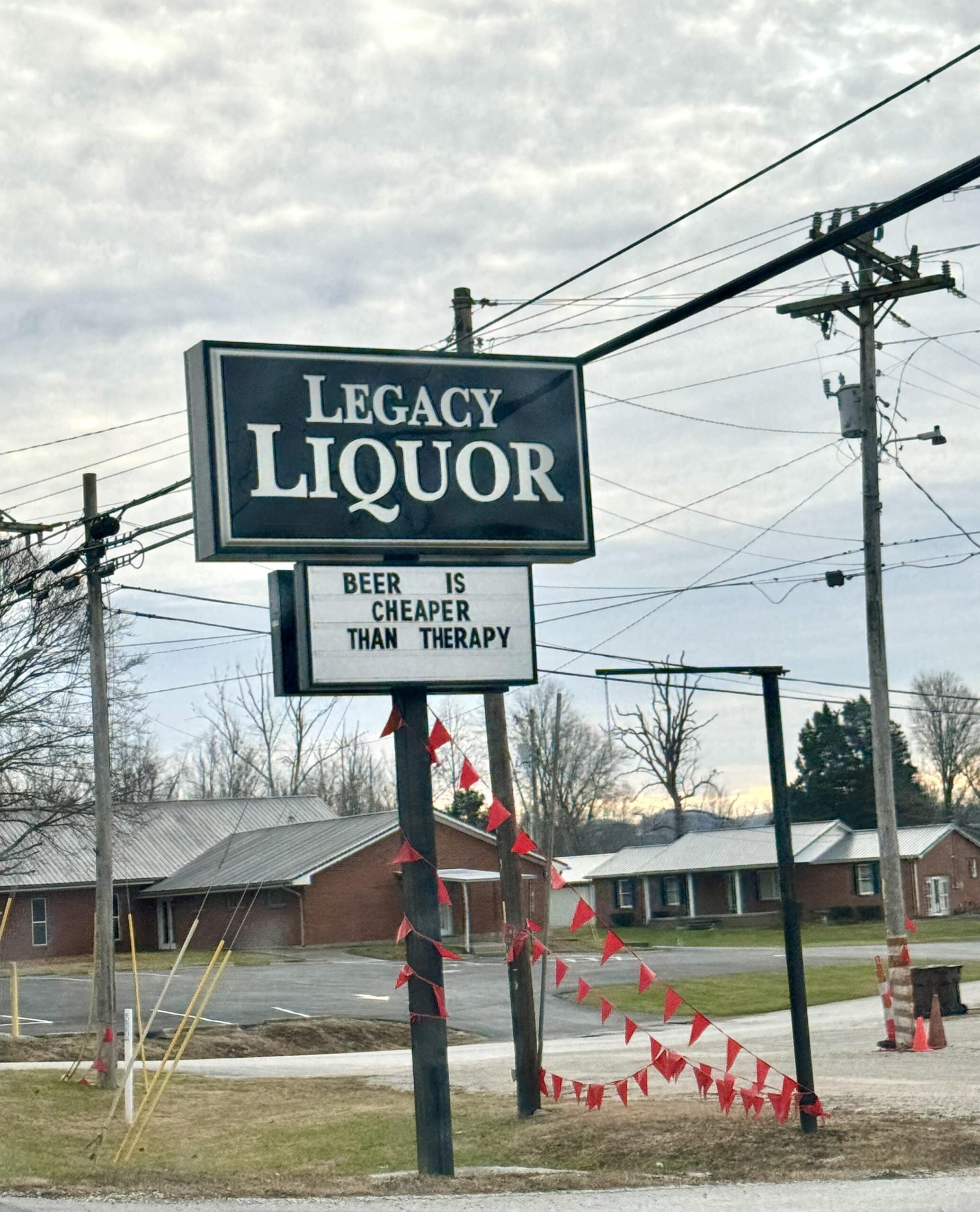 A sign for Legacy Liquor stands outside, with a message below reading, "BEER IS CHEAPER THAN THERAPY." Red flags and a few houses are visible in the background under a cloudy sky.