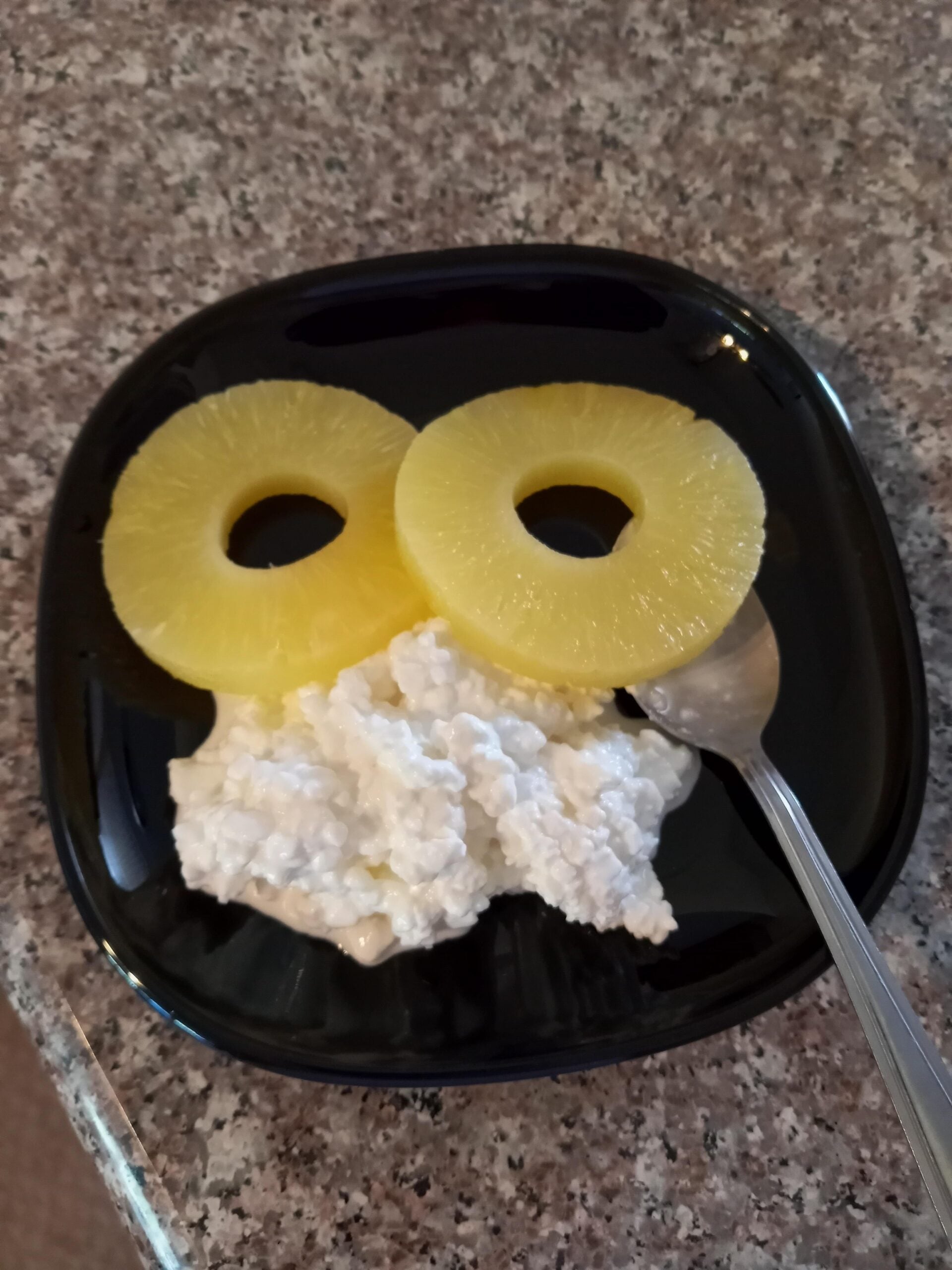 A black plate with two pineapple rings and a serving of cottage cheese, arranged to resemble a face, with a spoon resting on the side, on a speckled countertop.