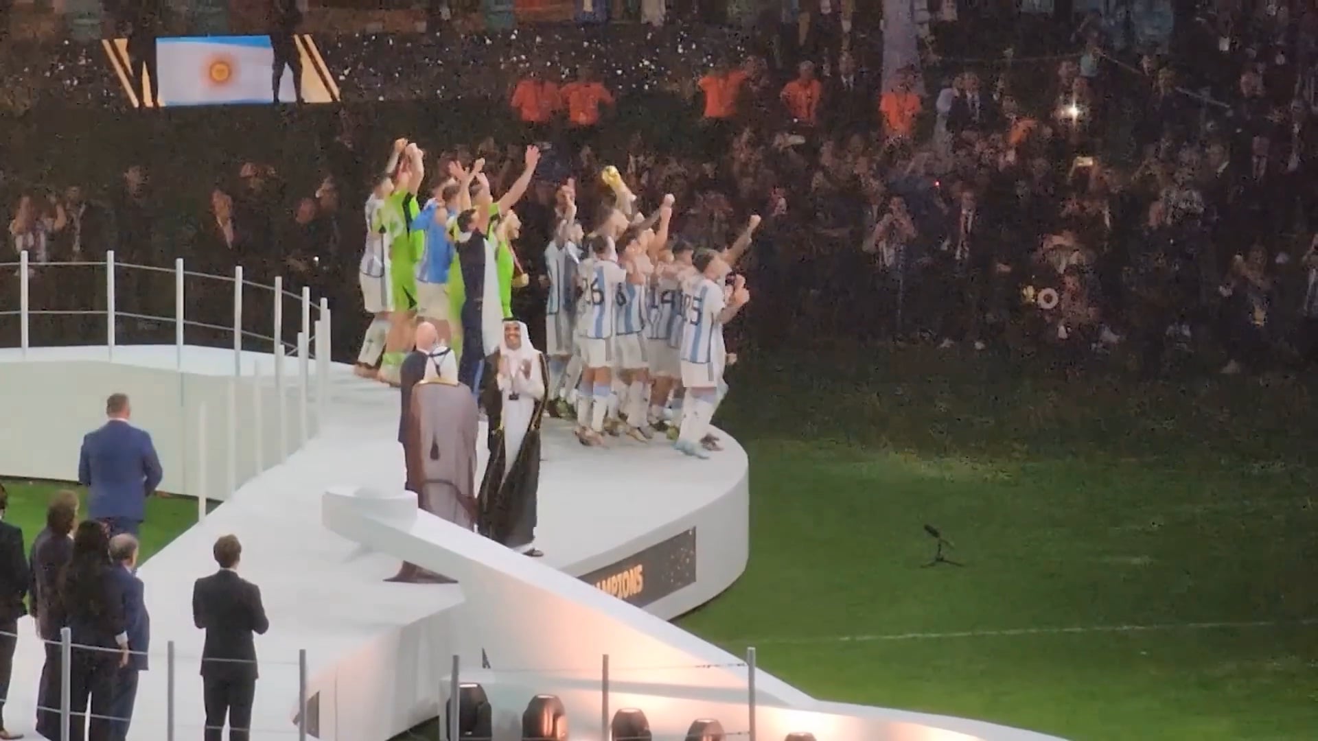 A soccer team in white and blue uniforms celebrates with a trophy on a podium, confetti falling as officials stand nearby and a crowd watches in the background.