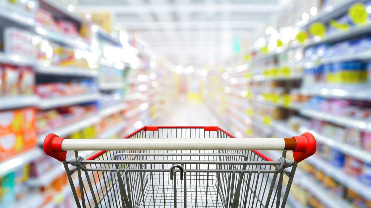 A shopping cart with a red handle is seen in focus inside a brightly lit grocery store aisle, with shelves on both sides stocked with various colorful products, blurred in the background.