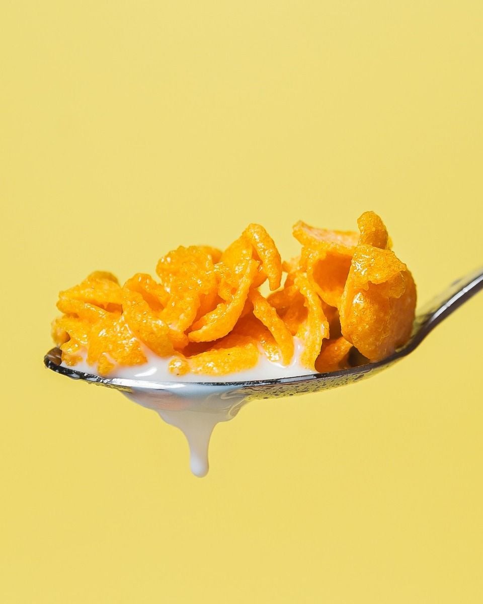 A close-up of a spoon holding cornflakes with milk dripping off the edge, set against a solid yellow background.