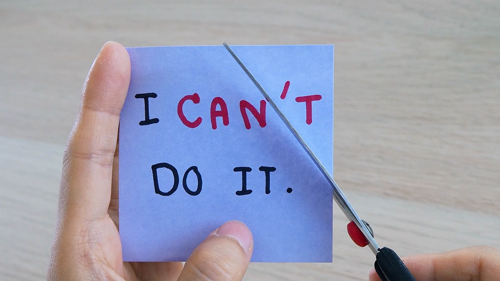 A hand holds a note reading "I CAN'T DO IT." while another hand uses scissors to cut between "CAN" and "T," changing the message to "I CAN DO IT.