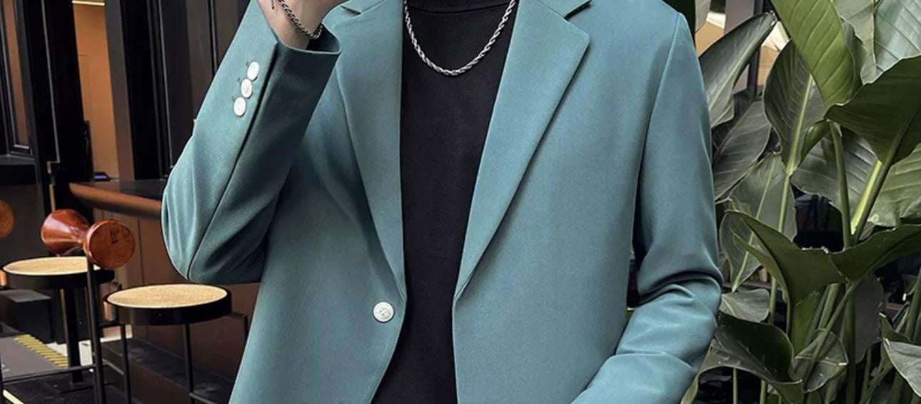 A person wearing a teal suit jacket over a black shirt and a silver chain necklace stands indoors near green plants, with part of their face out of frame.