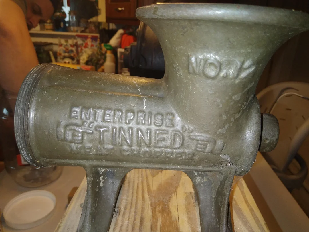 A close-up of an old metal meat grinder labeled “Enterprise Tinned” on its side, resting on a wooden surface with kitchen items and a person blurred in the background.