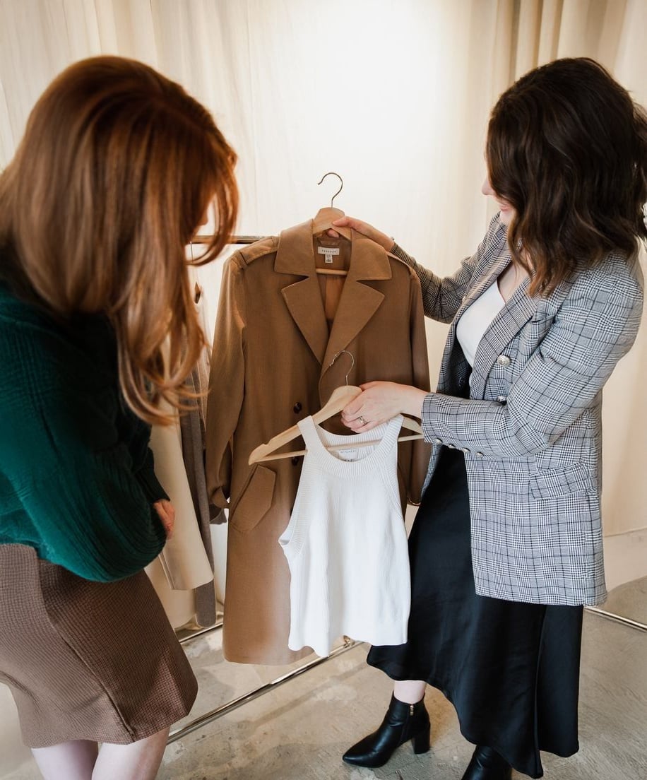Two women stand indoors examining clothing items on hangers; one holds a tan trench coat, and the other holds a white sleeveless top. Both appear to be discussing or choosing the outfits.