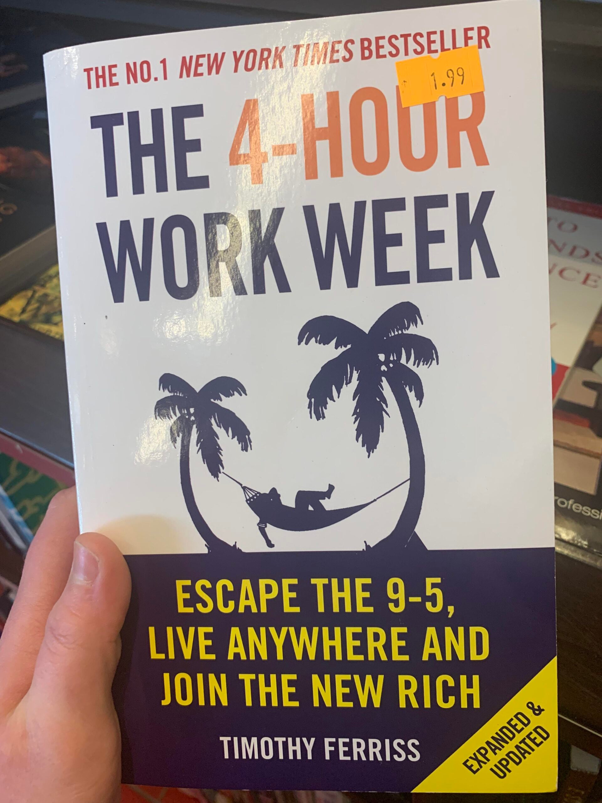 A hand holds the book "The 4-Hour Work Week" by Timothy Ferriss. The cover shows a hammock strung between two palm trees and features the text: "Escape the 9-5, live anywhere and join the new rich." A price sticker reads $1.99.