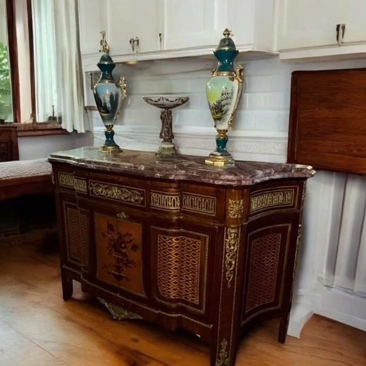 An ornate wooden cabinet with gold detailing and a marble top stands against a white wall, holding two decorative vases and a small pedestal dish in a bright, elegant room with wooden flooring.
