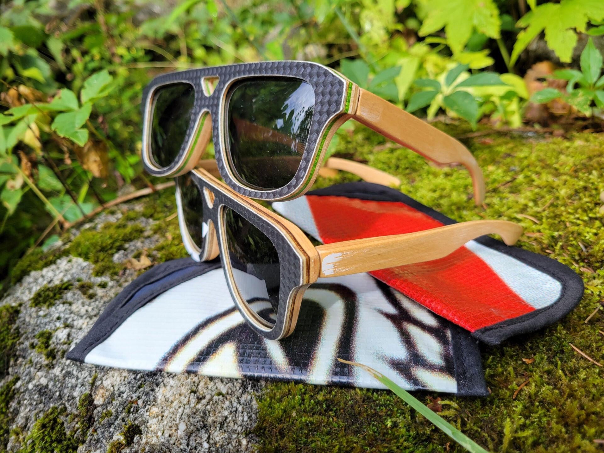 Two pairs of sunglasses with wood and carbon fiber frames rest on a mossy rock, with a soft case featuring a red and white design underneath. Green foliage surrounds the scene.