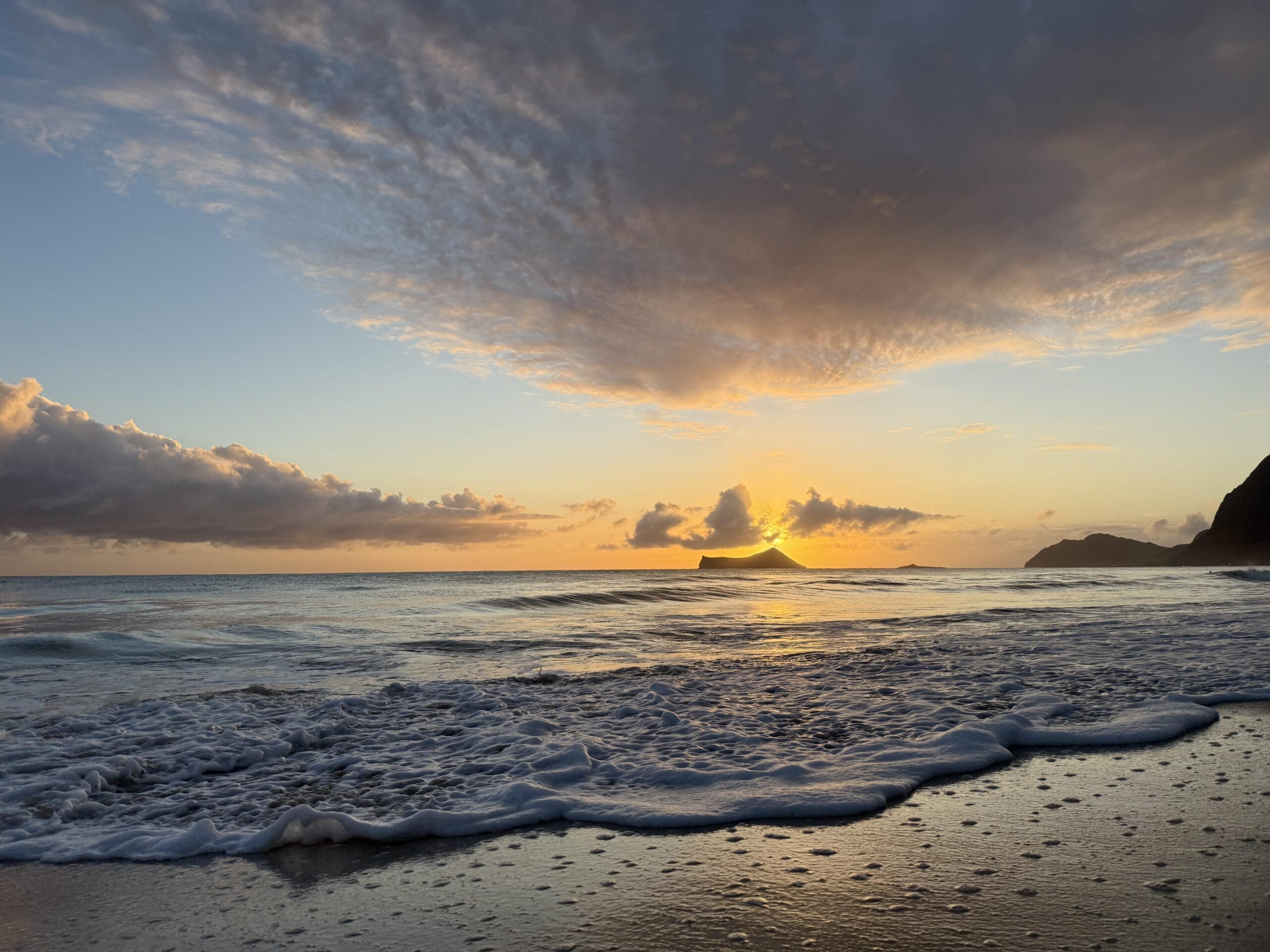 Sunset over the ocean with gentle waves washing onto the sandy shore, clouds scattered across the sky, and a silhouette of an island in the distance beneath a dramatic golden sky.