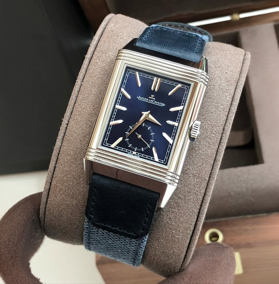 A rectangular luxury wristwatch with a blue dial, silver hour markers, and a blue leather strap is displayed on a brown padded holder. The brand name "Jaeger-LeCoultre" is visible on the dial.