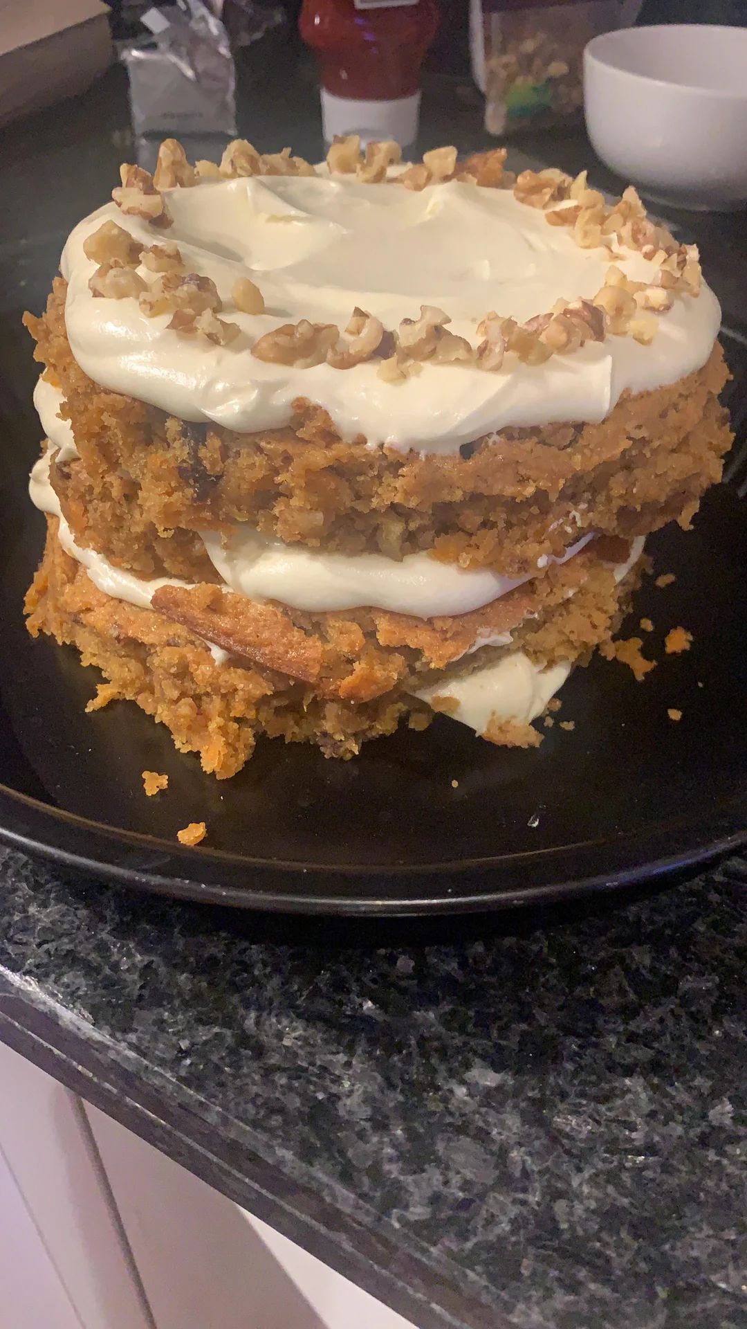 A homemade, two-layer carrot cake with cream cheese frosting and chopped walnuts on top, displayed on a black plate on a kitchen countertop.
